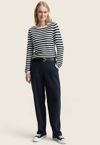 Chemise à manches longues à rayures bleues marines et blanches associée à un pantalon bleu marine ample et une ceinture noire. Le modèle porte des baskets noires et blanches.