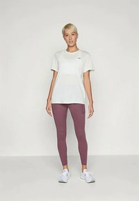 Bela kratka majica s kratkimi rokavi s potiskanem logotipom, v kombinaciji s prilegajočimi temno vijoličnimi leginami in belimi športnimi čevlji.