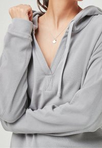 Světle šedá mikina s kapucí z texturované látky, s výstřihem do V, kapucí a dlouhými rukávy. Je vidět stříbrný náhrdelník s malým přívěskem.