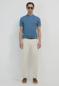 Polo shirt azzurro a maniche corte con tre bottoni, abbinato a pantaloni color crema e mocassini marroni. Il modello indossa occhiali da sole scuri.