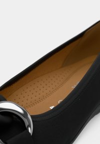 Svart skinnballettflat med avrundet tå, teksturert innfelt del og en metallisk detalj foran. Glatt innside med perforeringer.