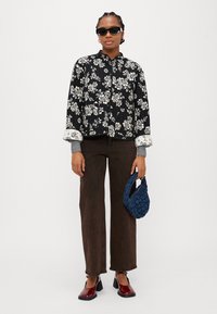 Giacca nera floreale con motivi bianchi, polsini grigi, pantaloni larghi marrone scuro, scarpe rosse lucide e borsa intrecciata blu navy.