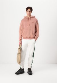 Sudadera rosa con capucha y bolsillo tipo canguro, combinada con pantalones de chándal blancos con franjas laterales verdes. La modelo sostiene un bolso color tostado y lleva zapatillas negras.