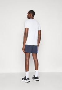 Λευκό αθλητικό μπλουζάκι κοντό μανίκι συνδυασμένο με ναυτικό σορτς με μπλε λεπτομέρεια, μαύρες αθλητικές κάλτσες και λευκές κάλτσες.