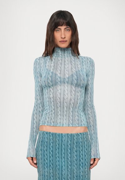 Femme portant un haut transparent à manches longues en tricot bleu à motifs torsadés et une jupe assortie, se tenant devant un fond blanc uni.