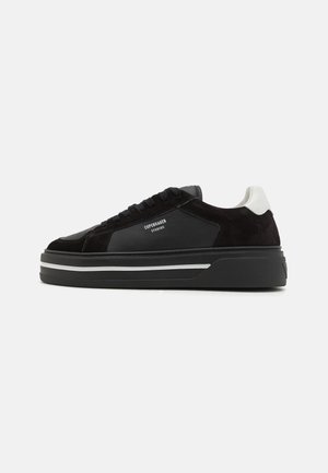 Svart lær- og semsket sneakers med en kraftig svart såle. Har et hvitt hælakkent og merkevaretekst på siden. Snøring.
