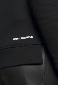Primer plano de una tela negra con el logotipo en blanco "KARL LAGERFELD" por encima de un dobladillo acanalado, acompañado de pantalones de cuero negros con una costura de bolsillo visible.