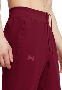 Pantalones deportivos burdeos con textura suave, cinturilla elástica, detalles de cordón y bolsillos laterales. Presenta un logotipo sutil en el muslo.