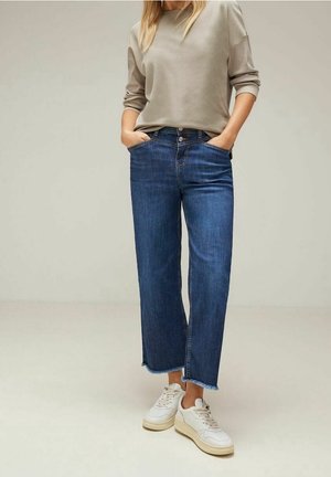 Kobieta ubrana w beżową bluzkę z długim rękawem, niebieskie jeansy o wysokim stanie z postrzępionymi końcami i białe trampki, stojąca z rękami w kieszeniach.