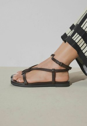 Fuß mit schwarzer flacher Sandale mit dünnen Riemen und Knöchelschnalle, kombiniert mit schwarz-weiß gestreiften, weit geschnittenen Hosen.