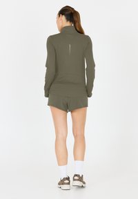 Olivgrünes Sportoberteil mit hohem Kragen, langen Ärmeln und strukturiertem Rückenpanel, kombiniert mit passenden Shorts und neutralen Sneakers.
