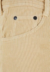 Tessuto in corda beige con rib legati verticali, dotato di una tasca con bottoni in metallo nero e cuciture a vista, che evidenziano la sua texture e il design.