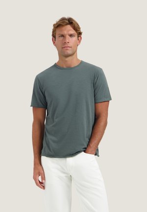 Man met kort blond haar, gekleed in een grijs T-shirt met korte mouwen en een witte broek, staand tegen een effen lichte achtergrond.