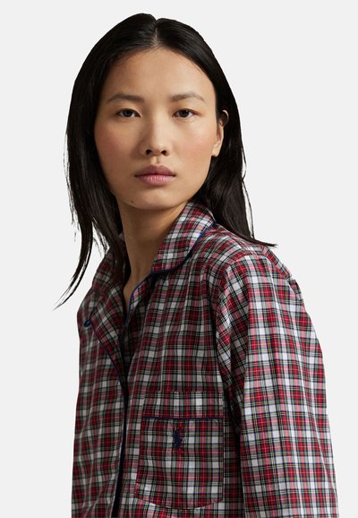 Mladá žena s dlouhými černými vlasy, oblečená v červeno-bílo-modré kostkované košili s knoflíky, stojící čelem k nám na jednoduchém světlém pozadí.