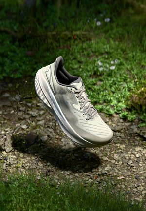 Chaussure de course gris clair avec semelle texturée flottant au-dessus d'un chemin rocheux avec de l'herbe et des plantes en arrière-plan.