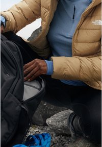 Doudoune couleur sable, chemise ajustée à manches longues bleue, sac à dos noir avec intérieur gris, leggings noirs et sandales bleues à motifs sur un sol rocheux.