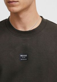 Dunkelgrüner Baumwollpullover mit Rundhalsausschnitt und einem rechteckigen blauen Etikett mit weißem Text auf der Brust. Strukturierte Stoffoberfläche.