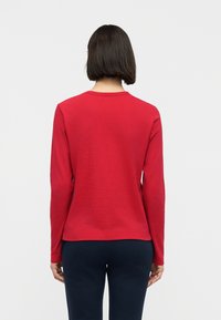 Γυναίκα με κοντά σκούρα μαλλιά που φοράει κόκκινο πουκάμισο μακρυμάνικο και σκούρο παντελόνι, όρθια με την πλάτη προς τα εμπρός, σε απλό φωτεινό φόντο.