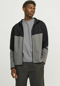 Giacca grigia e nera con zip e cappuccio, caratterizzata da un design bicolore e texture contrastanti, indossata sopra una t-shirt grigia.