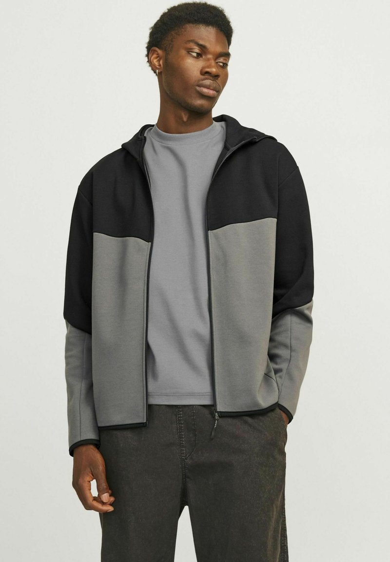 Giacca grigia e nera con zip e cappuccio, caratterizzata da un design bicolore e texture contrastanti, indossata sopra una t-shirt grigia.