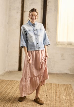 Femme souriante en veste en jean claire, jupe midi rose et chaussures marron sans lacets, debout sur un tapis tissé dans une pièce doucement éclairée.