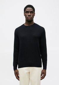 Pulover tricotat de culoare bleumarin, cu guler rotund, textură netedă, mâneci lungi, manșoane și tiv împăturit, având un logo discret pe piept. Purtați-l cu pantaloni bej.