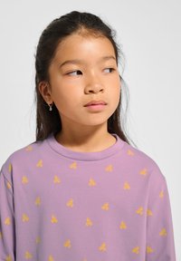 Junges Mädchen mit langen dunklen Haaren, das ein lila Shirt mit gelben blumenähnlichen Mustern trägt und nach rechts schaut, vor einem schlichten Hintergrund.