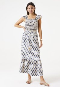 Maxi-jurk in crèmekleur met blauwe bladerenpatronen. Kenmerkt een smock-borstvoeding, gerimpelde mouwen en een uitlopende zoom met een trekkoorddetail.