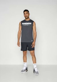 Siva brezrokavnika z napisom "RAIDERS Football" natisnjenim v belem, v kombinaciji s temnimi kratkimi hlačami in črno-bež športnimi čevlji.