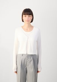 White cropped cardigan with a v-neck, long sleeves, and a single button closure, layered over a light top and paired with grey pinstriped trousers.