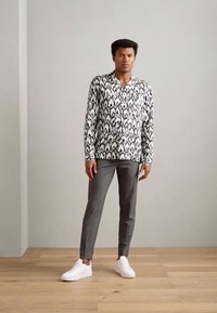 Srajca s črno-belimi vzorci, siva slim-fit hlače in bele superge. Gladka tekstura materiala, sodoben kroj in kontrastne barve.