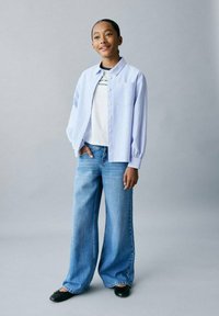 Cămașă cu dungi albastre deschise, cu butoni, purtată peste o bluză albă, asortată cu blugi evazați de denim albastru deschis și balerini negri, stând pe un fond gri.