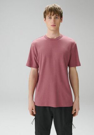 Jaunas vyras stoviniuoja veidu į priekį, dėvėdamas tekstūruotą dulksnos rožinės spalvos trumpomis rankovėmis palaidinę ir juodus krovinius kelnes, fone yra paprasta pilka siena.