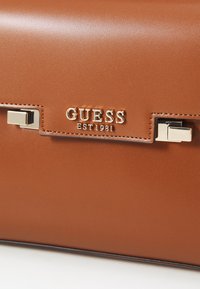 Borsa in pelle marrone con una texture liscia. Presenta una chiusura in metallo argentato e il logo impresso "GUESS EST 1981" in bella vista.