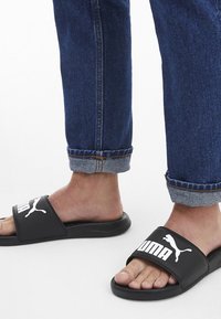 Sorte sutsko med hvide Puma-logoer på stroppen, lavet af syntetisk materiale, båret med opkrøllede blå denimjeans.