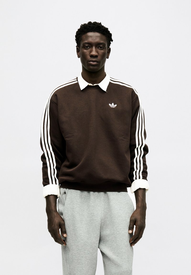 Giovane uomo in piedi, indossa una felpa marrone Adidas con strisce bianche sulle maniche e pantaloni della tuta grigio chiaro, sfondo neutro.