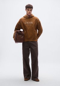 Brun hoodie med framficka och vit logotyp, kombinerad med mörkbruna vida byxor och bruna skor. Modellen håller en brun skinnväska.