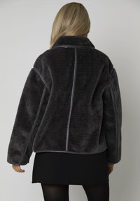 Femme aux longs cheveux blonds portant une veste en polaire gris foncé sur une jupe noire, se tenant de dos face à la caméra.