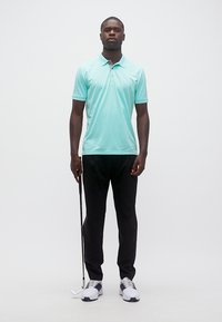 Άνδρας που στέκεται κρατώντας ένα μπαστούνι γκολφ στο δεξί του χέρι, φορώντας ανοιχτό μπλε πόλο, μαύρο παντελόνι και λευκά παπούτσια γκολφ πάνω σε ένα απλό φόντο.