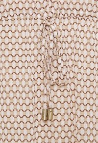 Beige tyg med ett strukturerat geometriskt mönster i brunt och krämfärgat. Inkluderar en dragsko med gyllene detaljer.
