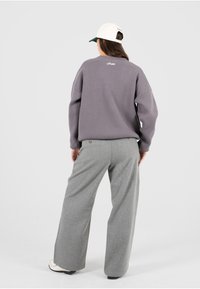 Pull en laine gris surdimensionné avec des poignets côtelés et un détail de logo, associé à un pantalon large gris clair et une casquette blanche avec une visière verte.