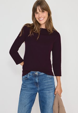 Burgunderfarbendes Langarmshirt mit Rundhalsausschnitt, kombiniert mit blauen Jeans. Das Shirt sitzt eng, hat eine glatte Textur und Dreiviertelärmel.
