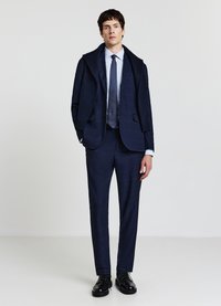 Marineblauer maßgeschneiderter Anzug mit strukturierter Oberfläche, bestehend aus einem Mantel mit Schalkragen, schlanken Hosen und einem weißen Hemd mit gepunkteter Krawatte. Schwarze Schnürschuhe.