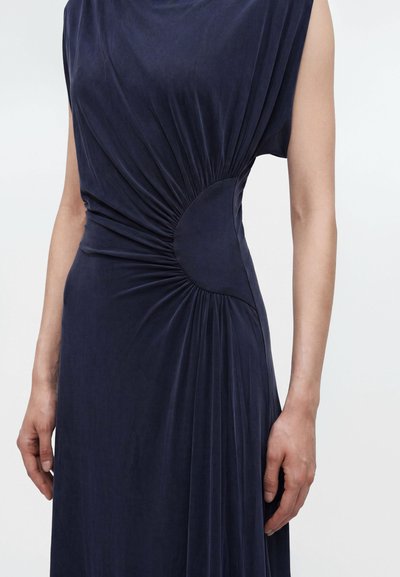 Personne portant une robe bleu foncé sans manches avec tissu froncé à la taille, créant un détail froncé circulaire d’un côté.