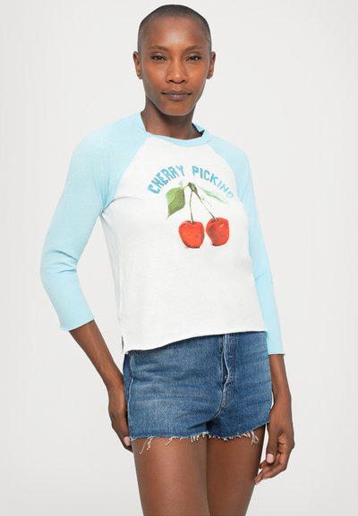 Woman wearing light blue and white long-sleeve shirt with cherries and "CHERRY PICKING" text, paired with high-waisted denim shorts.