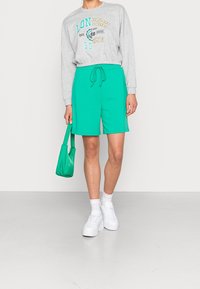 Grå sweatshirt med texten "LONYORK", tillsammans med ljusgröna shorts och vita sneakers. Håller en liten grön väska, står på en enkel yta.