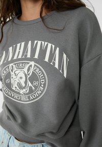 Grauer Sweatshirt mit einem runden Grafik eines französischen Bulldogs und dem Text "MANHATTAN" darüber in Weiß, geripptem Rundhalsausschnitt und leicht lockerer Passform.