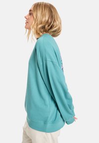 Tyrkysový oversized sweatshirt s kulatým výstřihem, spadlými rameny, žebrovanými manžetami a potiskem na zádech v růžové a fialové barvě. Měkká textura.