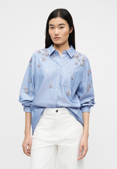 Γυναίκα που φοράει μπλε ριγέ πουκάμισο με λουλουδάτα κεντήματα και λευκό ψηλόμεσο παντελόνι, στέκεται μπροστά σε απλό φωτεινό φόντο.