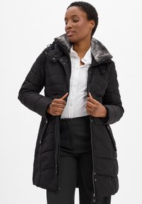 Zwarte gewatteerde winterjas met een gevoerd kraag van grijs bont en een voorliggende rits. Gekleed met een wit shirt en gestreepte broek.